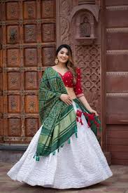 Woman in traditional lehenga with red blouse and green dupatta, accessorized with jhumkas, posing in front of an ornate wooden door — a perfect look for festive fashion, ethnic style, and wedding season trends.